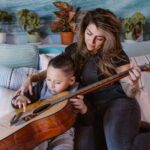 relaxed young woman playing guitar on bed with little son