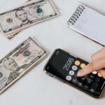 crop unrecognizable financier using calculator on smartphone near dollar banknotes
