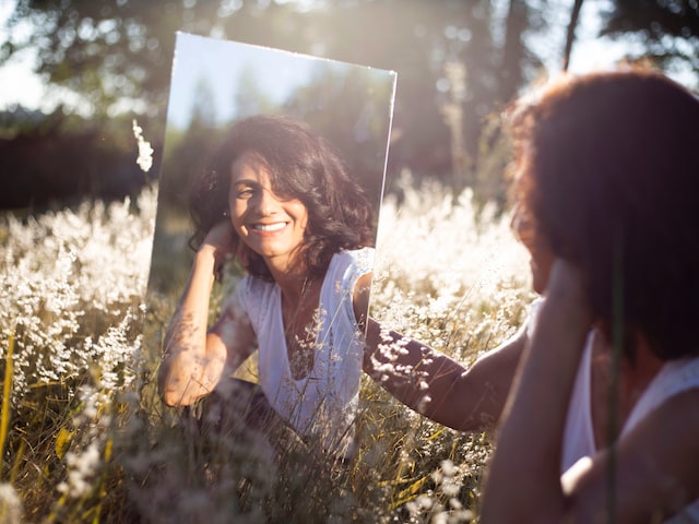 Woman looking at her reflection in a mirror smiling, while sitting in a field.