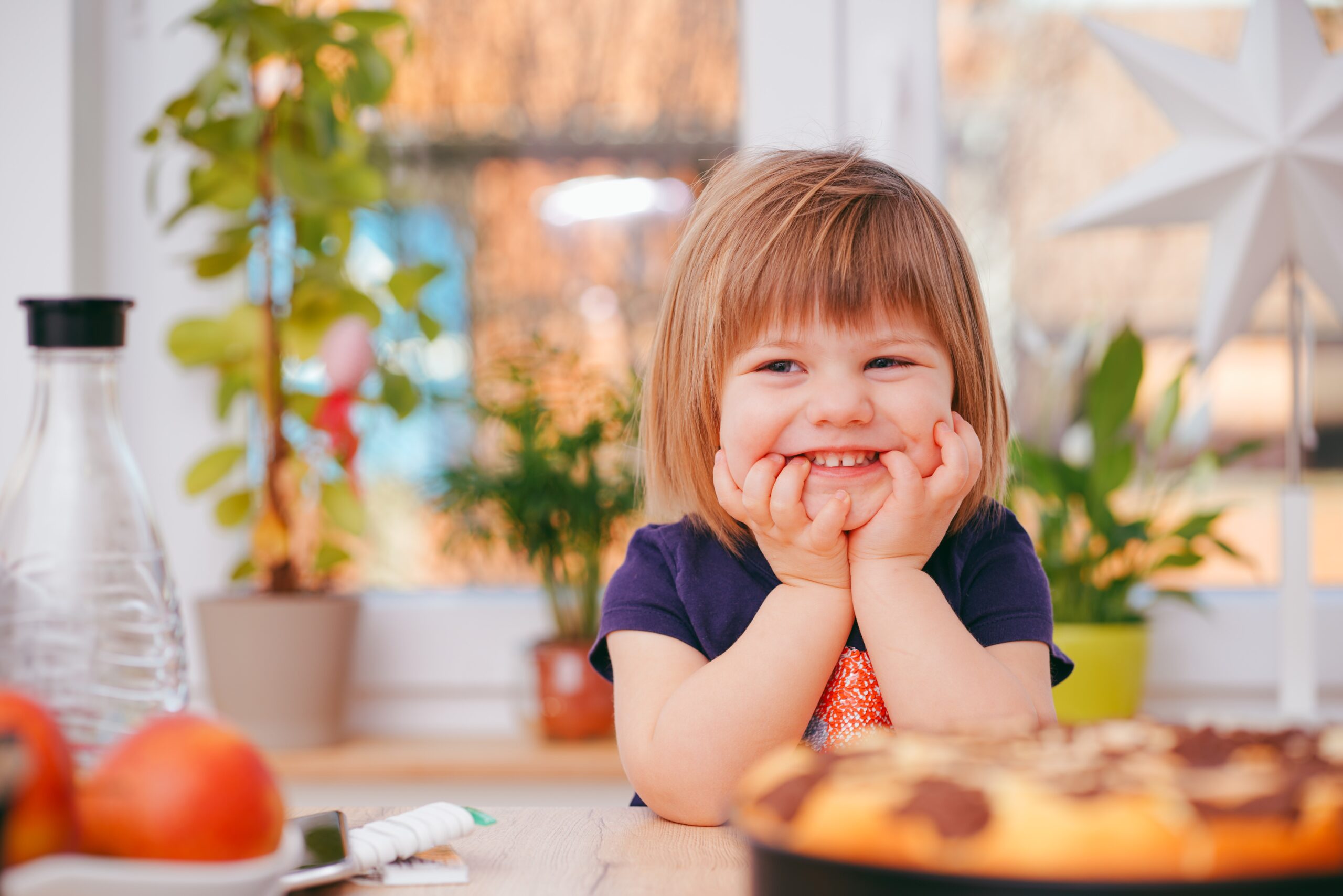 Toddler aged girl sitting at table with her elbows up and hands under her chin while smiling.