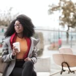 selective focus photo of woman in gray coat sitting on wooden chair holding a to go cup of coffee