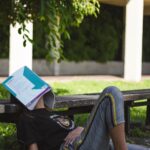 A student sitting on the ground with a textbook on their face, dozing off.