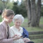 Younger and older woman sitting together on a bench outside while smiling.