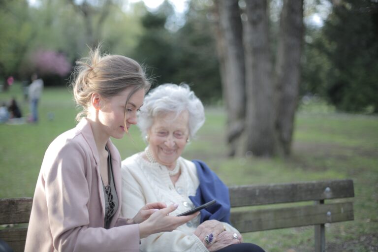 Younger and older woman sitting together on a bench outside while smiling.