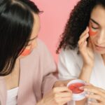 two women using red cosmetic pads and putting them on under eyes