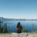 A person with long hair sitting on a rock looking at a lake.