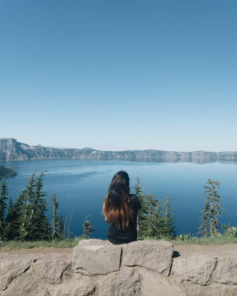 A person with long hair sitting on a rock looking at a lake.