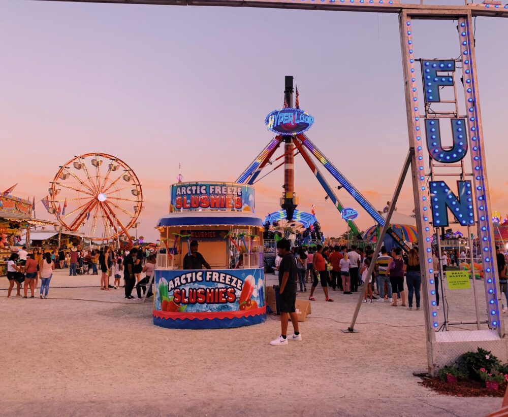 A wide shot of a fairly large crowd spread out among different attractions as a carnival or fair.