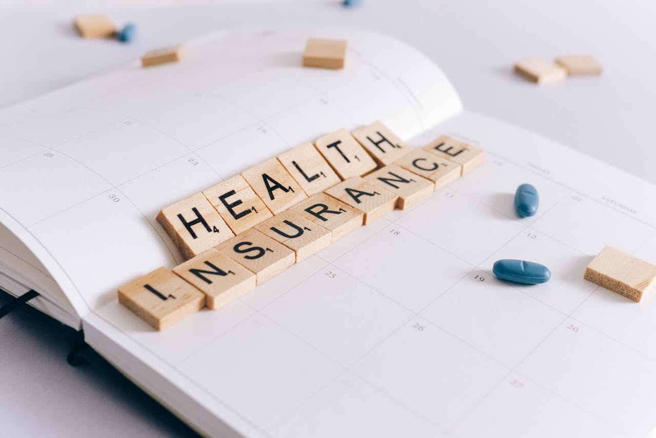 A series of Scrabble tiles arranged to spell "Health insurance" with pills scattered loosely nearby.