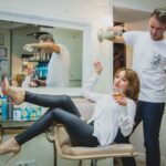 woman sitting on the salon chair while holding vodka glass and man at her back white spraying her hair