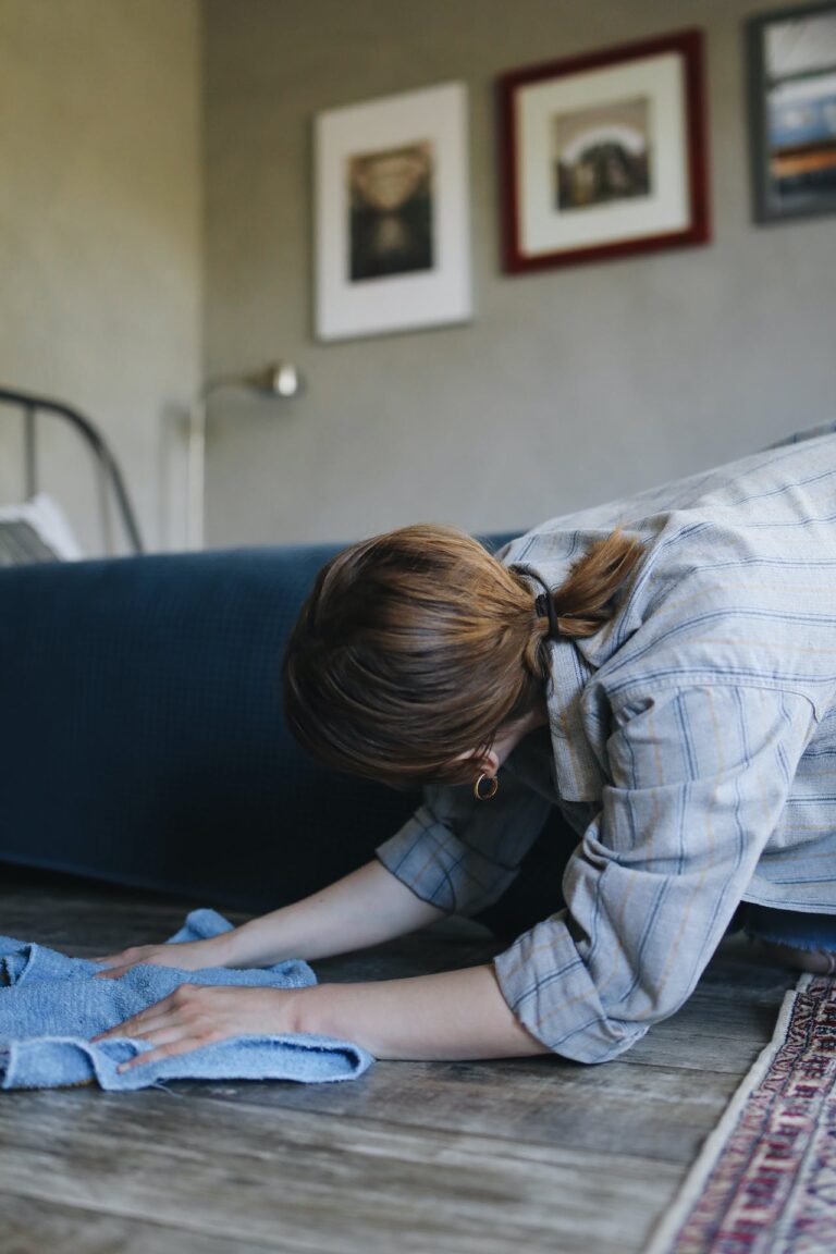 A person on hands and knees cleaning their floor while looking under a couch