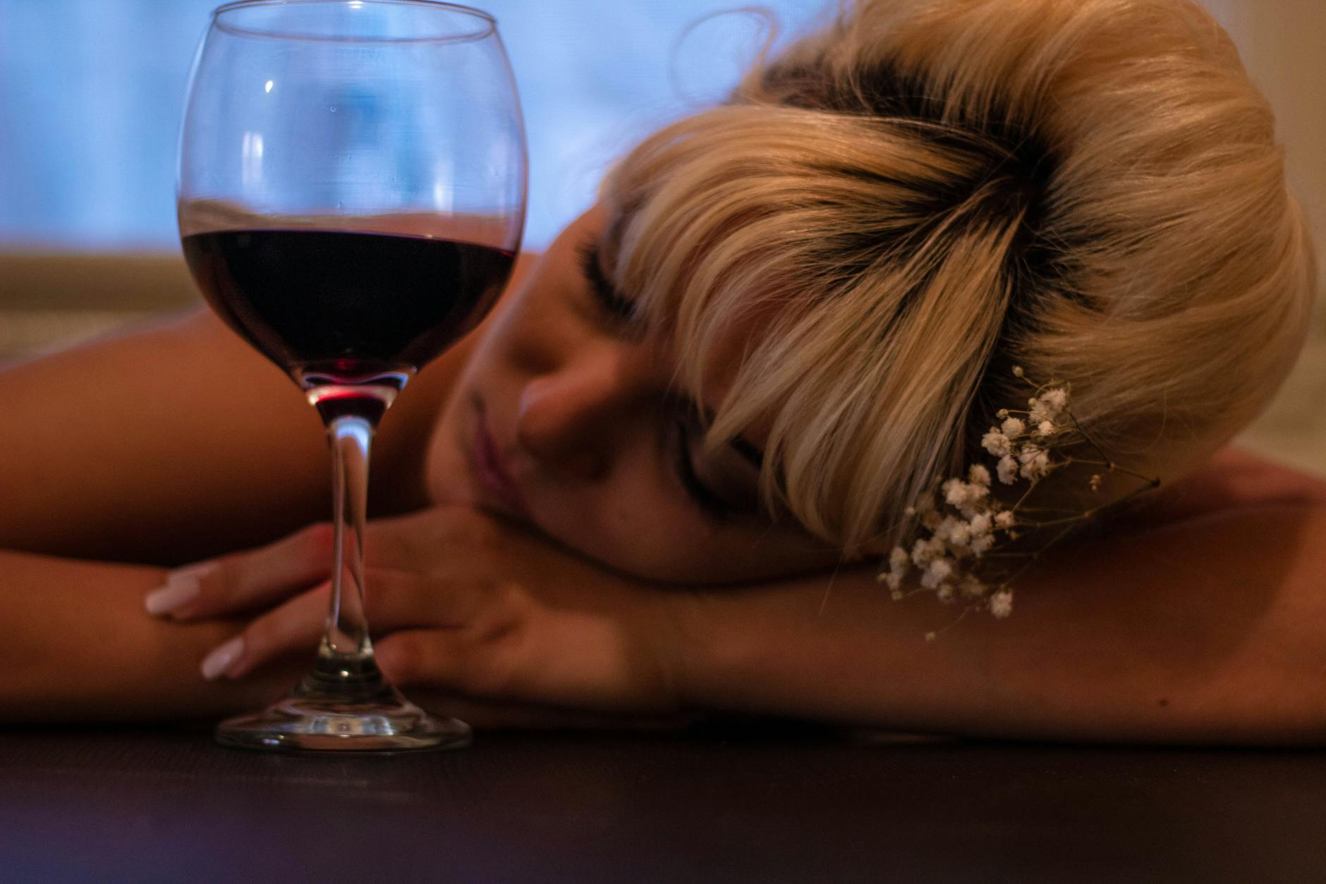 woman with white flower accent headdress leaning her head on table beside half filled wine glass