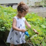 A young girl using a tiny watering can to water large collection of plants nearly as tall as her.