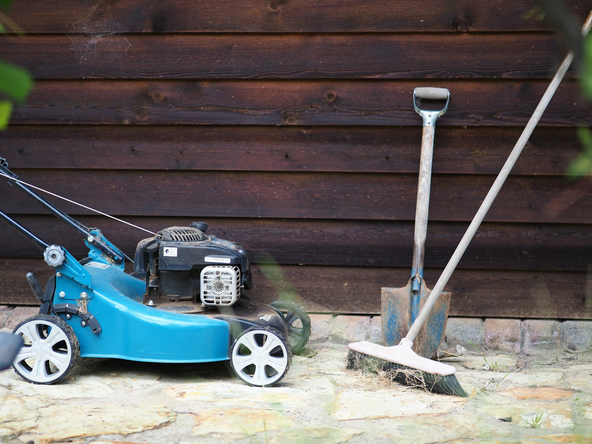 A lawn mower, a broom, and a shovel set against a shed.