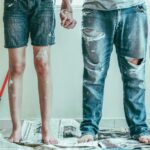 Two people, pictured from the waist down, holding painting tools in one hand and the other's hand in the other, standing in the middle of a room being painted.