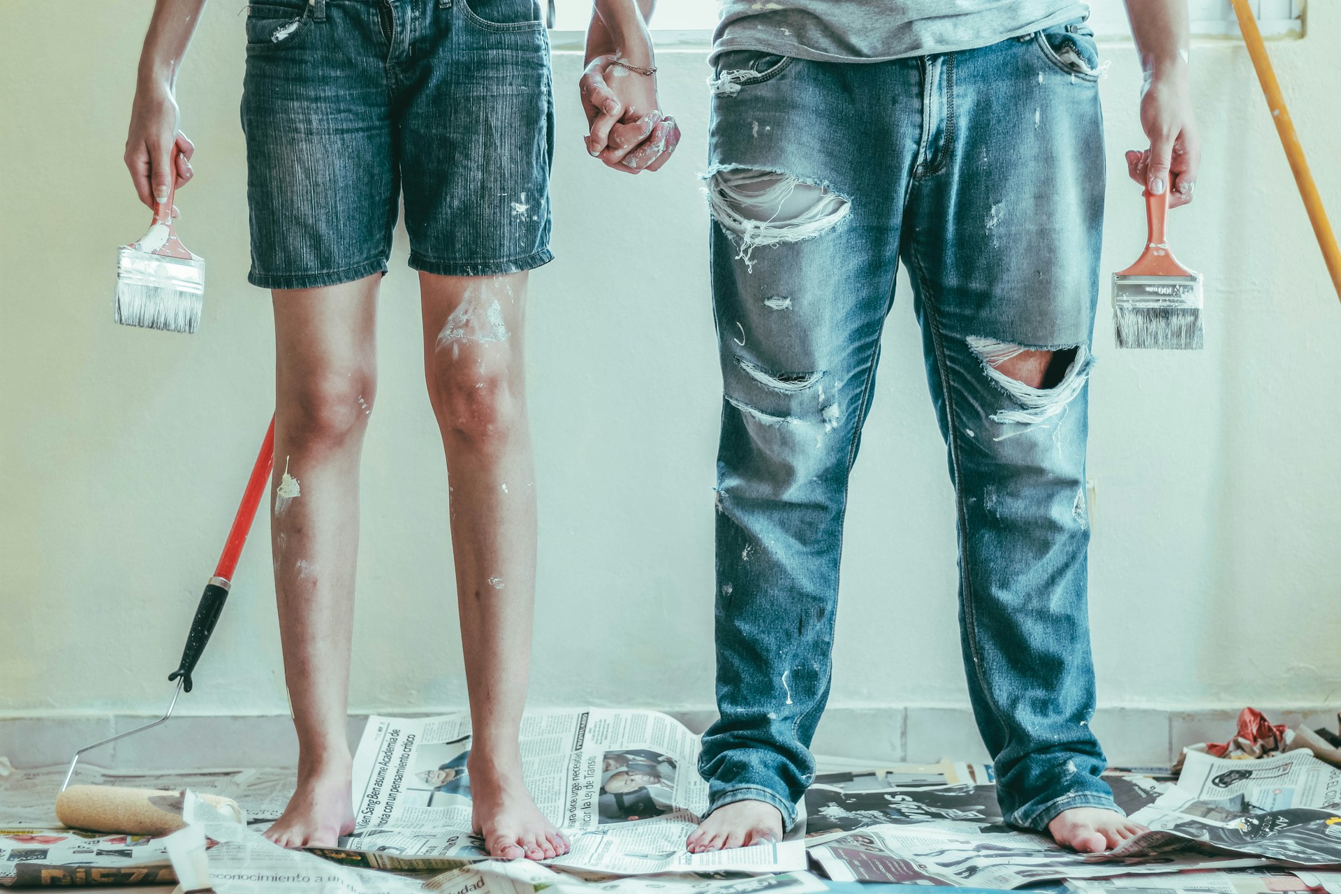 Two people, pictured from the waist down, holding painting tools in one hand and the other's hand in the other, standing in the middle of a room being painted.