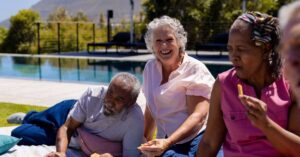 A diverse group of older adults sitting and smiling around a picnic blanket by a pool, eating from a charcuterie board.