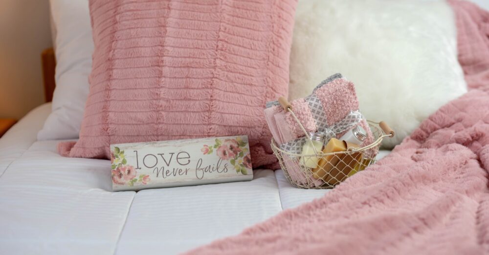 An extreme close-up showing a bed with white and pink pillows and blankets, plus a basket full of towels and soap.
