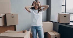 A woman wearing a t-shirt and jeans stands surrounded by cardboard moving boxes with her hands on her head appearing stressed.