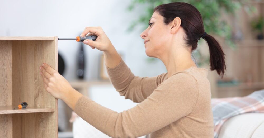 A woman with a ponytail is standing next to a tall wooden shelf. She's holding a screwdriver up against the side of it.