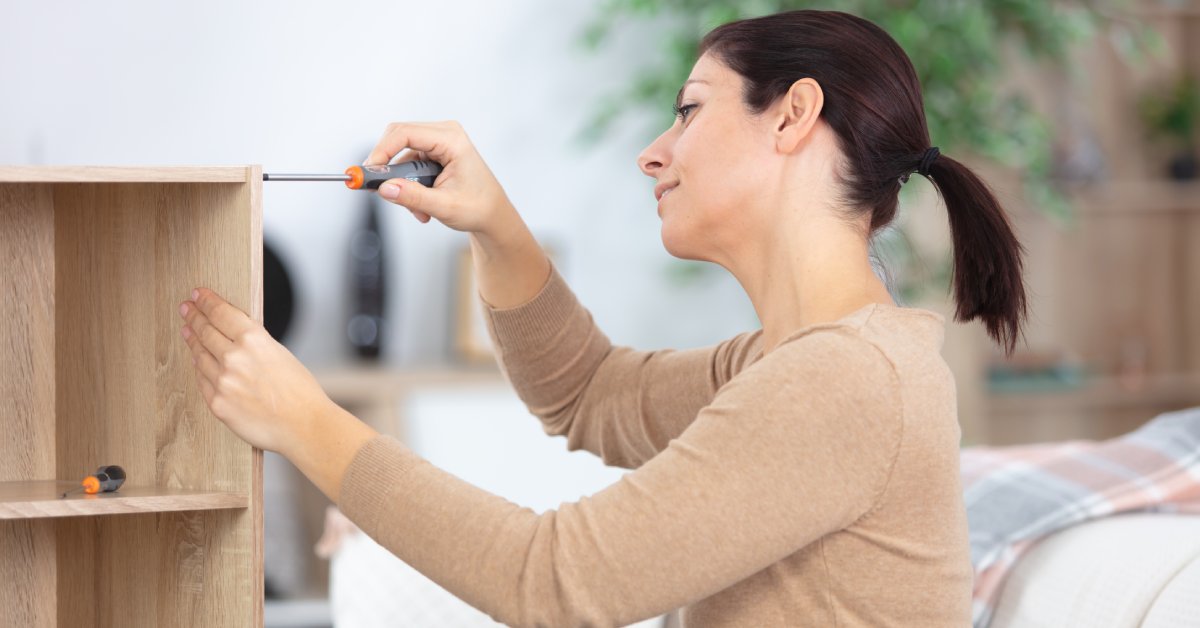 A woman with a ponytail is standing next to a tall wooden shelf. She's holding a screwdriver up against the side of it.