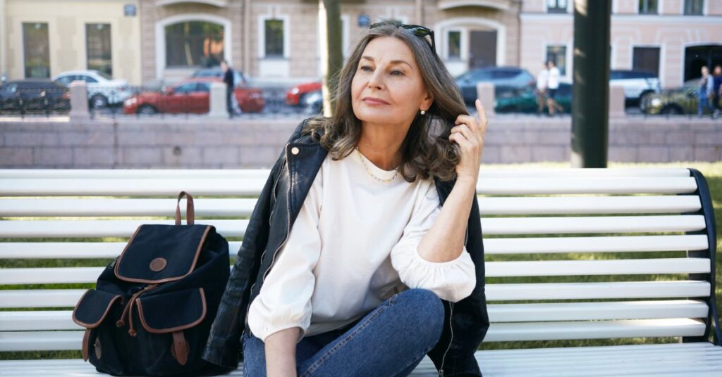 A confident, stylish middle-aged woman sits on a white park bench with one leg crossed, brushing her hair back from her face.