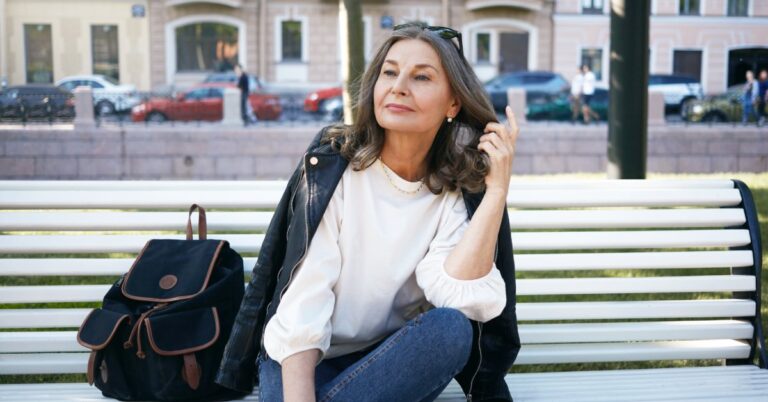 A confident, stylish middle-aged woman sits on a white park bench with one leg crossed, brushing her hair back from her face.