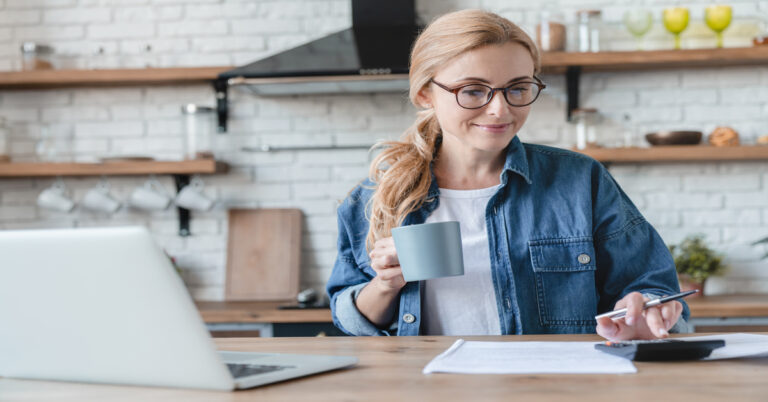 A mature blonde woman is calculating money in her kitchen, drinking coffee and working on her laptop.