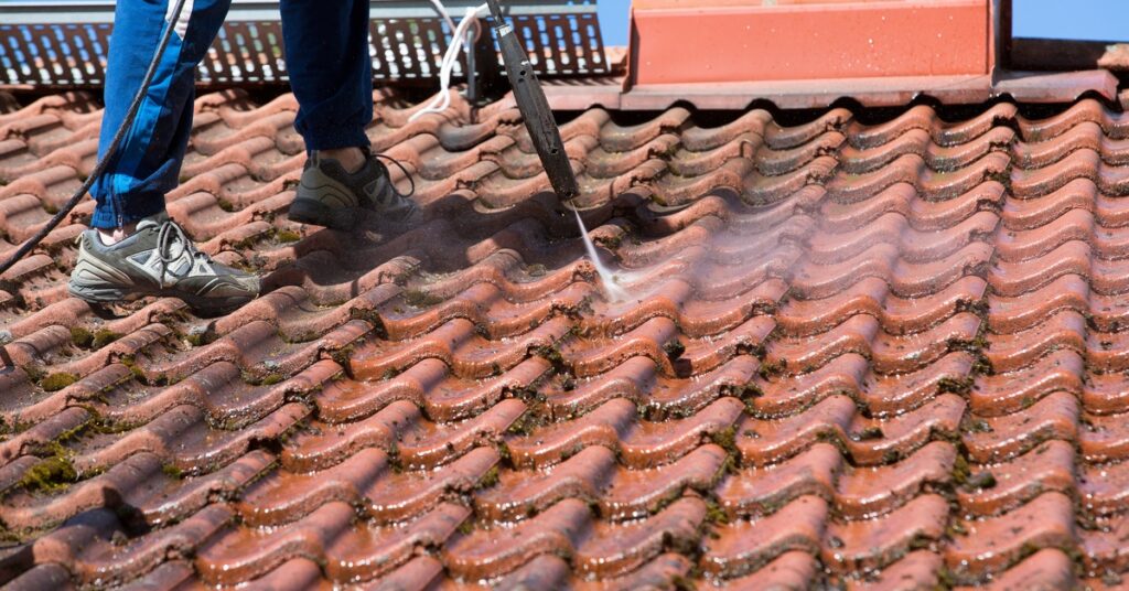 Water sprays from a pressure washer as a person cleans dirt, moss, and mold from a red architectural roof.