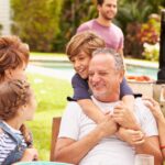 Smiling children and adults are gathered at a backyard table for a meal on a sunny day. More adults stand at an open grill.
