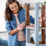 A woman wearing a blue denim jacket and red gloves sands down a light blue wooden chair with a sponge.
