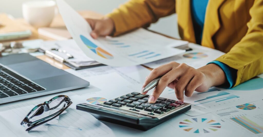 A businesswoman wearing an orange blazer sitting at a desk in front of financial documents with a calculator.