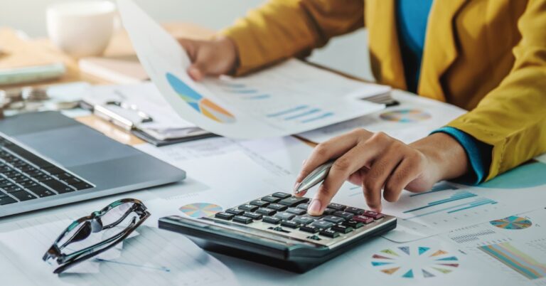 A businesswoman wearing an orange blazer sitting at a desk in front of financial documents with a calculator.