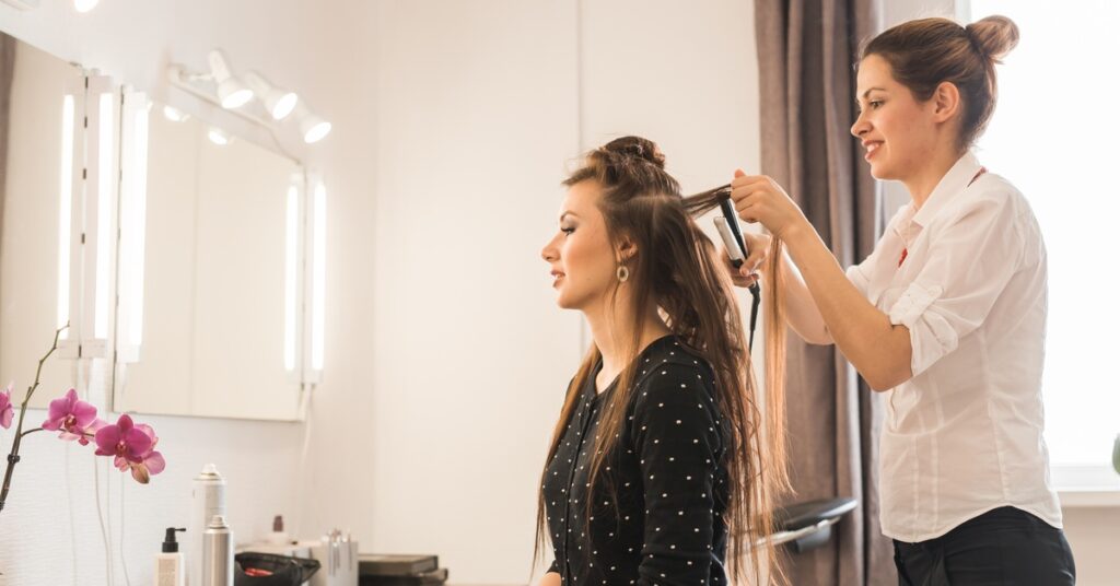 A woman wearing a white shirt uses a hair straightener to style another woman's hair inside a salon.