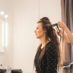 A woman wearing a white shirt uses a hair straightener to style another woman's hair inside a salon.