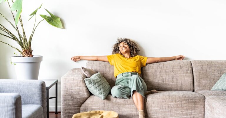 A smiling young woman in a yellow shirt and green pants relaxing on a tan couch next to a tall potted plant.