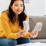 A smiling, long-haired woman in a yellow sweater sits on a gray couch with blue pillows, counting money.