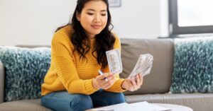 A smiling, long-haired woman in a yellow sweater sits on a gray couch with blue pillows, counting money.