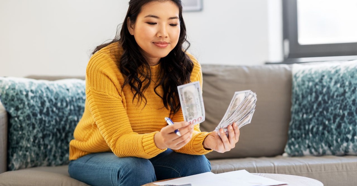 A smiling, long-haired woman in a yellow sweater sits on a gray couch with blue pillows, counting money.