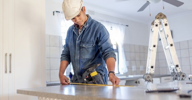 A home contractor wearing a denim shirt and a white hard hat uses a tape measure on a kitchen countertop.