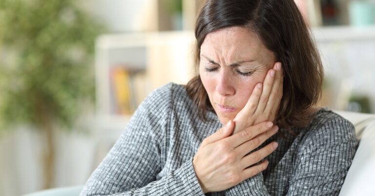 A woman wearing a gray sweater, sitting on a white couch, holds her hand up to her face while expressing pain.