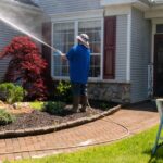 A man in a hat, blue shirt, and boots power washes the gray siding of a house with burgundy shutters.