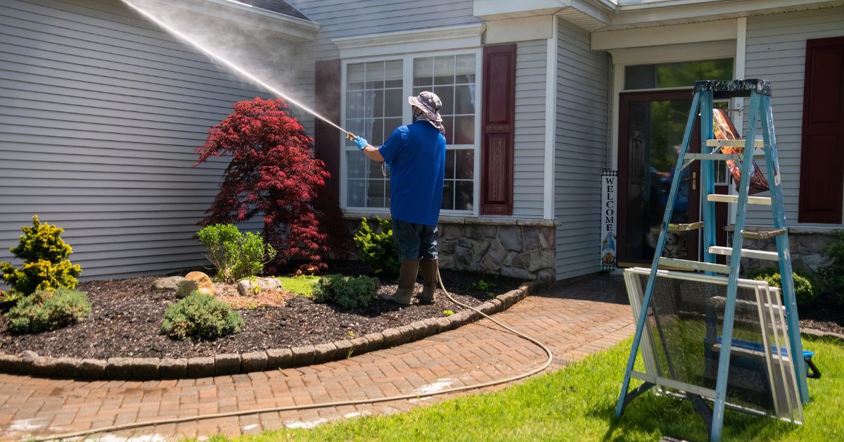 A man in a hat, blue shirt, and boots power washes the gray siding of a house with burgundy shutters.
