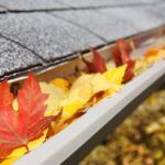 A silver gray rain gutter is filled orange and yellow autumn leaves, sitting next to a dark gray stone roof.