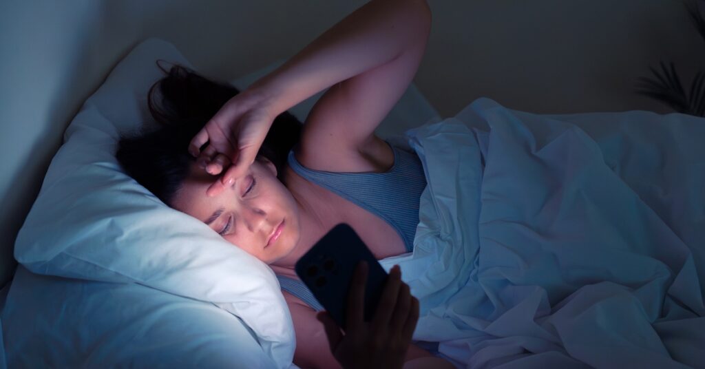 A woman lies in bed, squinting at her phone in the dark. The light from the screen illuminates her face.
