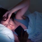 A woman lies in bed, squinting at her phone in the dark. The light from the screen illuminates her face.