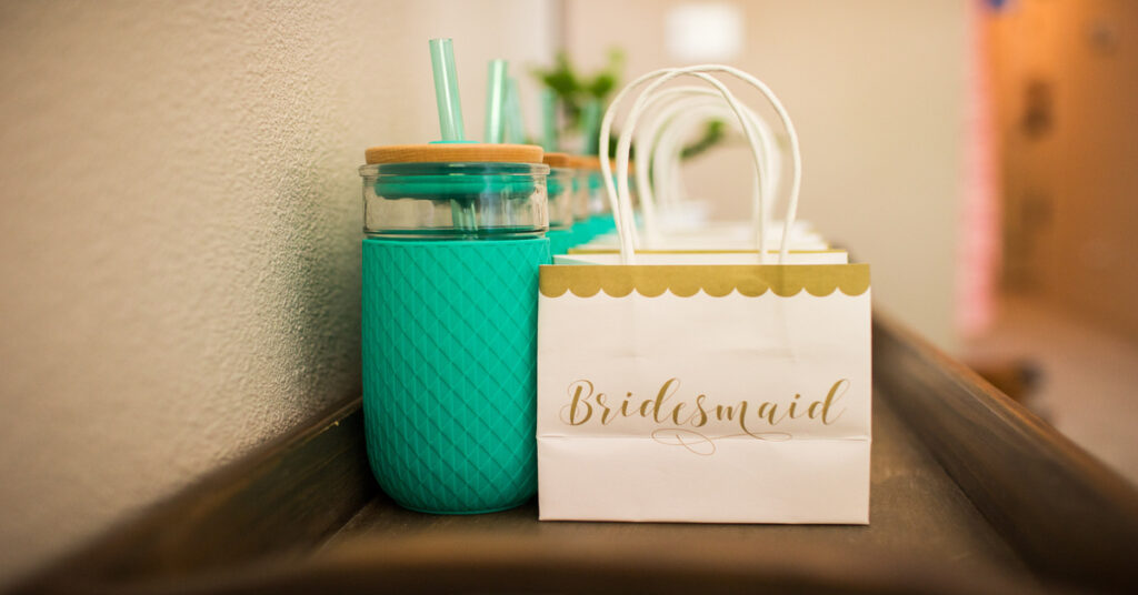 A row of white and gold gift bags labeled "Bridesmaid" sit next to a row of matching turquoise water bottles.