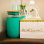 A row of white and gold gift bags labeled "Bridesmaid" sit next to a row of matching turquoise water bottles.
