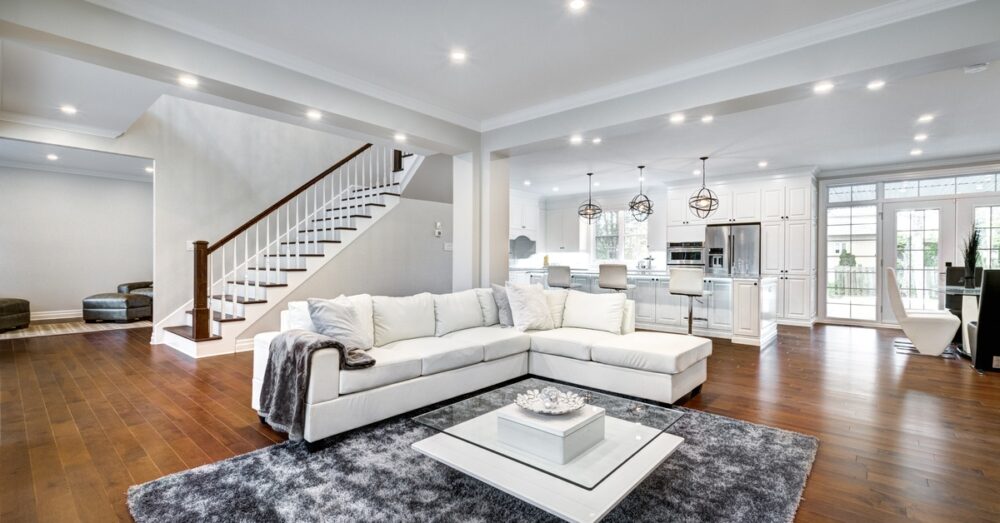 A large room with a white sectional couch and a grey couch next to a staircase with a kitchen in the background.