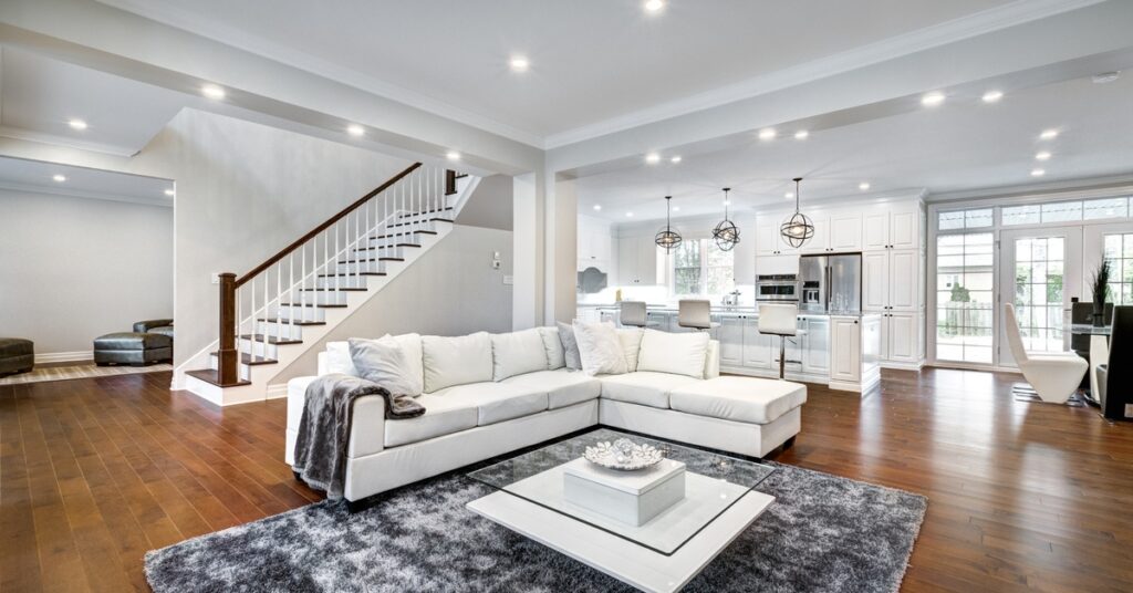 A large room with a white sectional couch and a grey couch next to a staircase with a kitchen in the background.